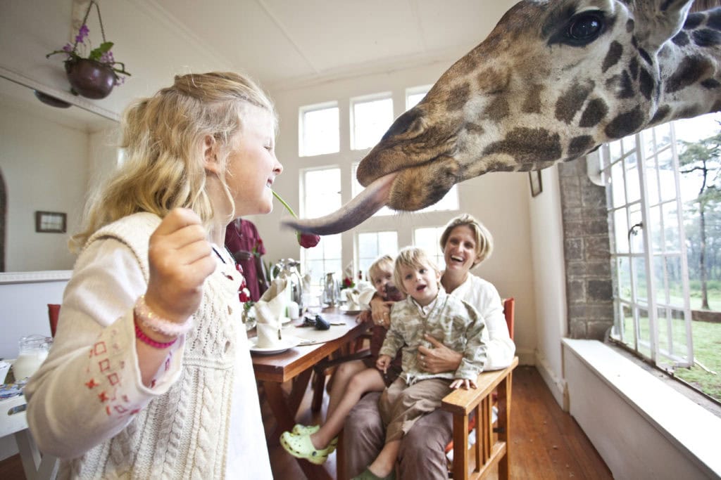 Image of a family with 3 young kids enjoying time with the giraffes at Giraffe Manor in Nairobi, Kenya. The daughter in the foreground smiles in delight as a giraffe extends its tongue | Planet Africa Safaris | Blog